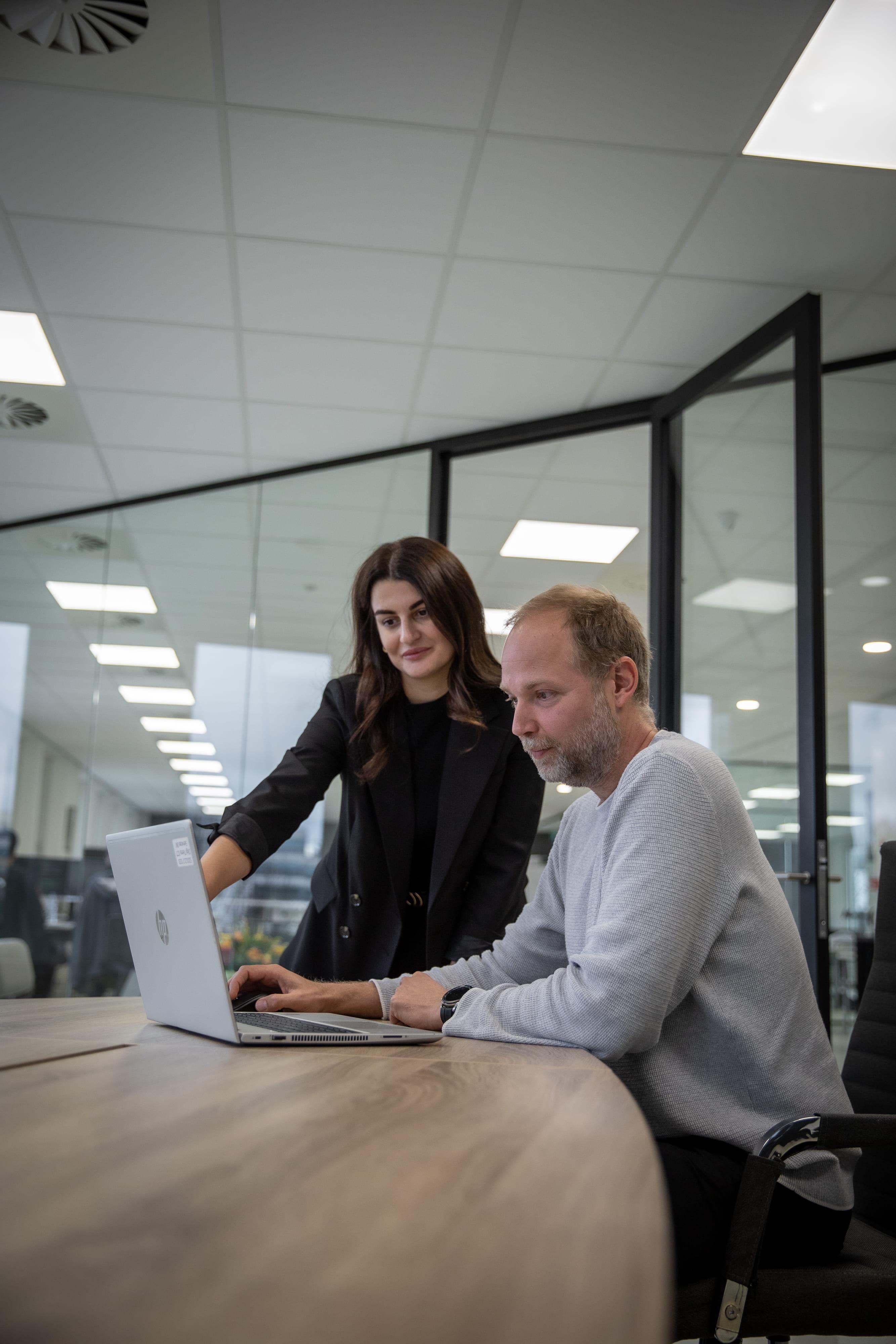 Een klant zit aan tafel met een laptop, terwijl een werknemer staand meekijkt en wijst naar de laptop.